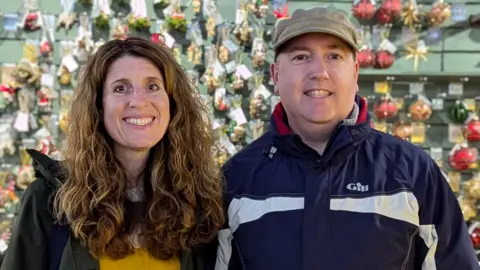 TBC Wesley Thorne and his wife Toni are smiling and standing in front of a wall covered in Christmas decorations.