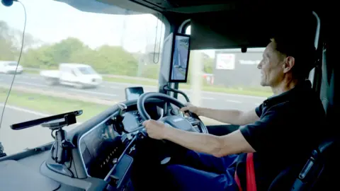 Criminals buy haulage firms to steal lorryloads of goods, BBC finds A lorry driver pictured inside his cab, sitting at the steering wheel, as he drives along a dual carriageway, with the view outside of passing vehicles slightly blurred as his lorry moves along