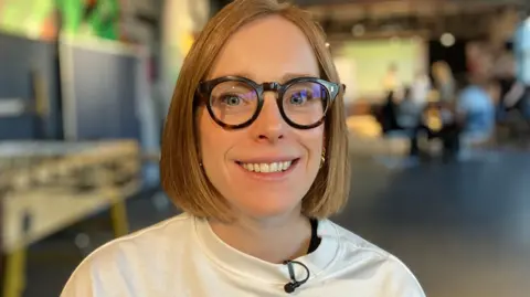 A white woman with a ginger bob wearing black glasses smiles at the camera. She's in a room where group coaching of young people is happening in the background. She's wearing a white sweater. 