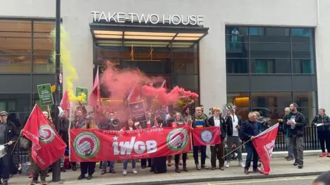 IWGB IWGB members holding a red banner emblazoned with the union's logo are shown picketing outside Take-Two House, the company's London HQ. People are shown holding signs, flags and megaphones as red and yellow smoke rises from smoke grenades.