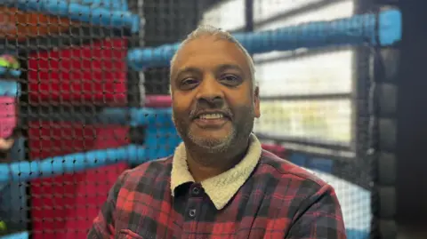 Errol Mittoo is standing in front of a play area. He is smiling at the camera and is wearing a checked jacket with a fur collar.