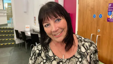 BBC A woman wearing a black and white top, with dark brown hair, standing inside a community centre - behind her are tables and chairs, and a cupboard.