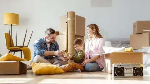 Getty Images A couple with a young boy sitting on the floor in a room surrounded by boxes that they are unpacking. The man and the boy are both holding and looking at a bronze coloured jug