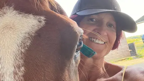 Sue - a woman smiling while wearing a baseball cap - stands next to a horse. The horse is closer to the camera, with only it's eye and forehead visible beside her.