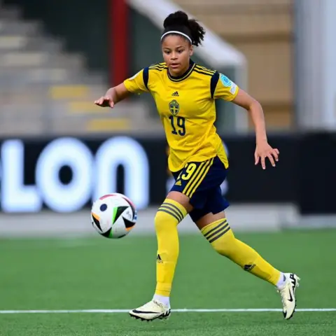 Getty Images Felicia Schröder playing football. She is wearing a yellow and black Sweden kit with the number 19. She is looking at the ball and controlling it while running.
