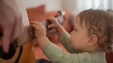 Getty Images A toddler reaches up to strum the strings of a guitar, being held by an adult
