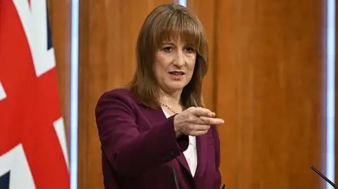 Getty Images Chancellor Rachel Reeves wears a plum coloured suit and points to a journalist while stood at a podium in the media briefing room of 9 Downing Street