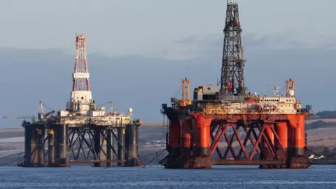 PA Media Two oil metal oil platforms, one red and the other black, in a flat blue sea, with low hills in the background. 