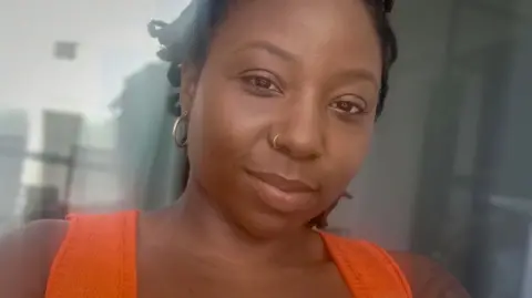 Serena Milius A woman with dark hair and a nose ring, wearing an orange top, smiles at the camera in a selfie