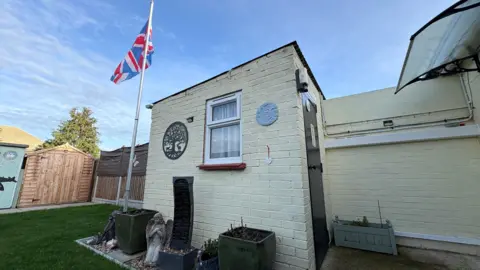 Ben Schofield/BBC A brick built shed, with cream walls, in a garden. We are looking at the side wall of the shed, which has a small window in it, through which we can see a net curtain. A black door into the shed is just visible in the front wall to the right. There is an area of grass to the left of the image and behind the shed. A flag pole also stands behind the shed, from which the Union Flag is flying. There are several garden ornaments including plant pots, a winged angel statue and a fountain water feature.