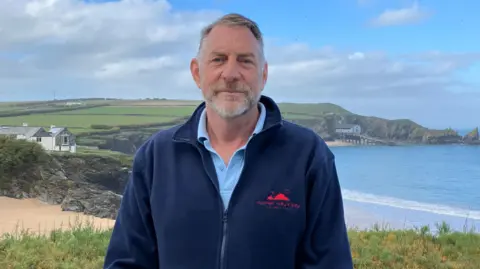 A man with great hair and beard wearing a dark blue fleece and light blue polo shirt with the sea and beach behind him. 