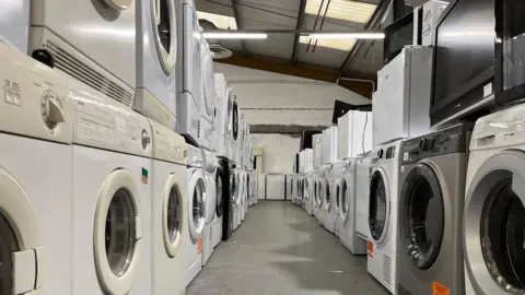 Looking down two rows of washing machines stacked in a warehouse. They are all different as they are second-hand. 