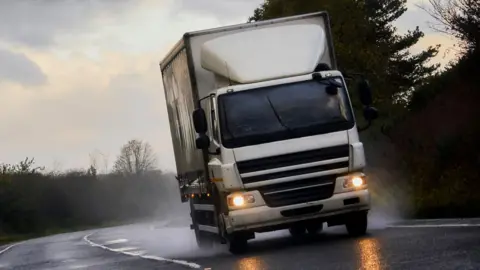 Getty Images A stock image showing a white HGV driving around a corner of a UK road, producing spray from recent rain. The daylight is fading and the lorry has its lights on.