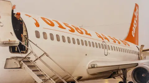 EasyJet Stelios stands at the top of the plane's steps waving at the camera. He wears an orange Easyjet baseball cap. The side of the plane has the Luton Airport phone number emblazoned in orange against a white plane. Is says 01582 445566. The tail of the plane is orange and has easyjet written in white.
