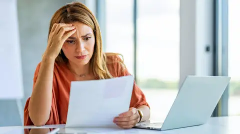 Getty Images/ljubaphoto A woman in a loose but smart top looks at papers whilst sitting in front of an open laptop. She is frowning slightly.