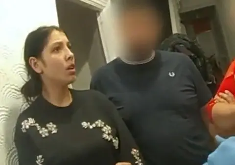 Met Police A woman with dark hair in a low bun is standing in her hallway, wearing a black jumper with white flowers on. She looks shocked.