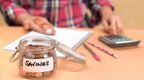 Getty Images Jar of coins with sticker saying "savings" in the foreground, with a hand, coins, calculator, pen, pencil and paperwork behind.