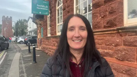Devon to see 1,300 more children get free school meals Sonia Duggan wearing a burgundy corduroy top with a black padded jacket and standing in front of the Family Hub building in Paignton