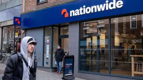 Getty Images Outside a Nationwide branch with the building and its blue name sign on in the background, a man walking out of the door of the branch and another man in a grey hoodie walking past in the foreground.