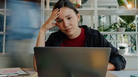 Getty Images A young woman wearing a dark blue shirt and a red t-shirt looks concerned as she looks at her laptop. She has her right hand on the side of her head.