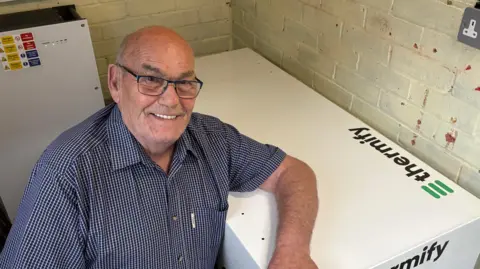 Ben Schofield/BBC Terrence Bridges kneeling down and smiling on the left of the frame, while leaning an arm over a large white object with "Thermify" written on it, which is on the right of the image. Terry is wearing a short-sleeved blue-checked shirt, which is open at the neck, and black-rimmed rectangular glasses. He is inside a small room, with brick walls. The bricks have been painted a cream colour. An unused plug socket is just visible in the top right of frame. Behind Terrence is another piece of electrical equipment with a white front, several warning stickers and two green lights.