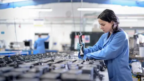 Getty Images A young woman wearing a blue boilersuit uses a tool on components sitting on a benchtop.