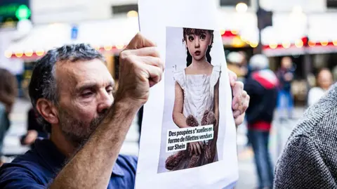 Getty Images A man holds up a poster with an image of a child-like sex doll with a caption in French criticising e-commerce giant Shein for selling the toys. The caption translates in English to "sex dolls shaped like little girls sold on Shein."