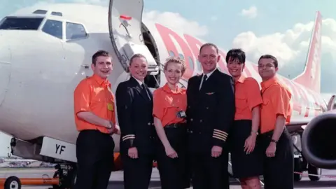 ITV Six crew members including a man and woman wearing pilot's uniforms stand in a group outside an Easyjet plane. Four of them wear bright orange short sleeved shirts and black trousers or skirts. They are all smiling.