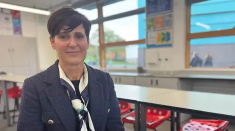 Mortgages and AI to be added to the curriculum in English schools Becky Francis is seated at a table in a classroom wearing a dark textured jacket and a patterned scarf. The room has white walls, large windows letting in natural light, and posters with educational content on the wall. There are red plastic chairs with holes in the seat arranged around white tables.