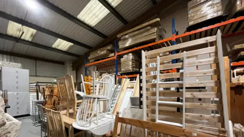 A picture of a warehouse with second hand tables, chairs and beds stacked on one another. In the background, carboard boxes with flat pack furniture which are seconds or returns from companies.