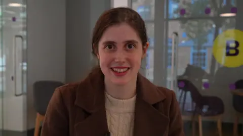 Katie Livings wearing a brown coat and cream cable knitted jumper with her dark hair tied back looking and smiling at the camera