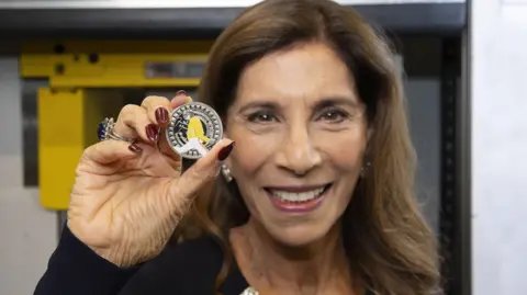 The Royal Mint Kashmira Bulsara smiles as she holds the Freddie Mercury coin. She has long brown hair and brown eyes. The coin is silver and has an image of Freddie Mercury on the front, mid performance, with his head titled back. The only colour on the coin is his yellow jacket and white trousers. 