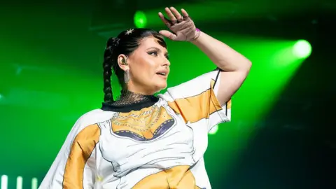 Getty Images Nelly Furtado performs on-stage, illuminated from behind by a green spotlight. She holds her left hand to her forehead, as if trying to make out something in the distance. She wears a white t-shirt printed with a picture of a female torso in a white vest-top and wearing several silver chains.
