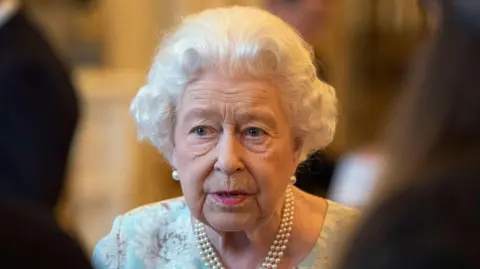 Getty Images A file photo of Queen Elizabeth at an October 2019 reception in Buckingham Palace, wearing a string of pearls, pearl earrings and a floral dress in pale blue and white with accents of taupe.