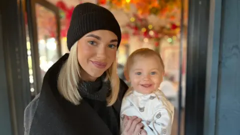An image of Lauren Harper pictured with Reuben, aged nine months, at a cafe in Eastbourne. Lauren is wearing a grey and black coat alongside a black hat. She has blonde hair. Reuben is wearing a white babygrow with bears. Both are smiling.