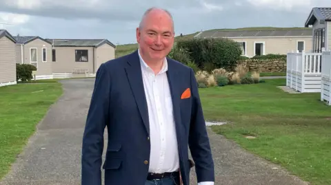 BBC A man with thinning grey hair wearing a white shirt and dark blue blazer standing on a wide path surrounded by static caravans. 