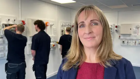 Lindsey Young, wearing a red jumper with a blue coat over the top, looks at the camera with a neutral expression. She has long blonde hair down to over her shoulders. In the background are three young men all dressed in black trousers and polo shirts working on electrical circuitry