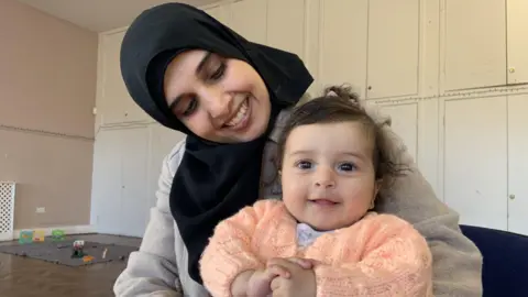 A woman wearing a black hijab is sitting down and smiling and holding a baby girl. The baby has short black curly hair, is wearing a peach coloured cardigan and is smiling
