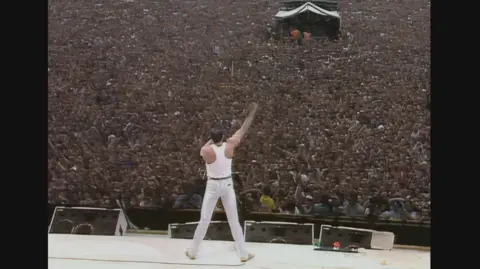 Band Aid Licensing Ltd. Freddie Mercury stands on the stage at Live Aid facing the packed audience. We see him from behind in a wide stance, he is dressed all in white - trainers, jeans, tank top. He holds the microphone to his mouth and his right arm is raised in a fist. 