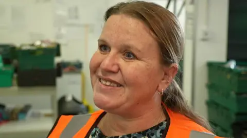 BBC Close up of Lisa Parkinson who stands in the warehouse in front of green food crates. She is wearing an orange high vis vest and has long fair hair tied back in a ponytail