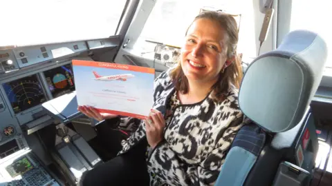 Cathy Adams Cathy sits in the cockpit of an Easyjet plane holding a certificate that is orange and blue. It says CONGRATULATIONS at the top. She is smiling at the camera and wears glasses on her head, and a black and white jacket and black trousers.
