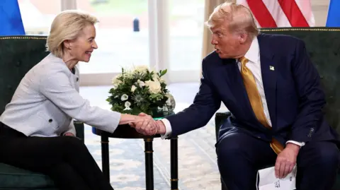 Reuters Von der Leyen, in a white cropped jacketa nd black pants reaches her hand in front of a side table with a white flower arrangement to grip the hand of Trump, who is in a blue suit and gold tie and holds papers in his other hand