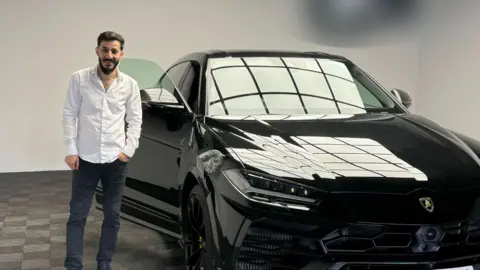 TikTok Benjamin Mustata, a man with dark hair and a beard, wearing a white shirt and dark trousers, smiles as he stands next to a black Lamborghini SUV with the driver's door open. The picture is taken indoors, in what appears to be a showroom