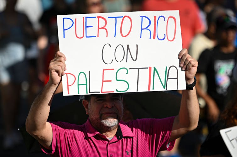 How both remix what it means to be Boricua A man in a red shirt, standing amid a crowd, holds a sign that says, in Spanish, 'Puerto Rico with Palestine.'