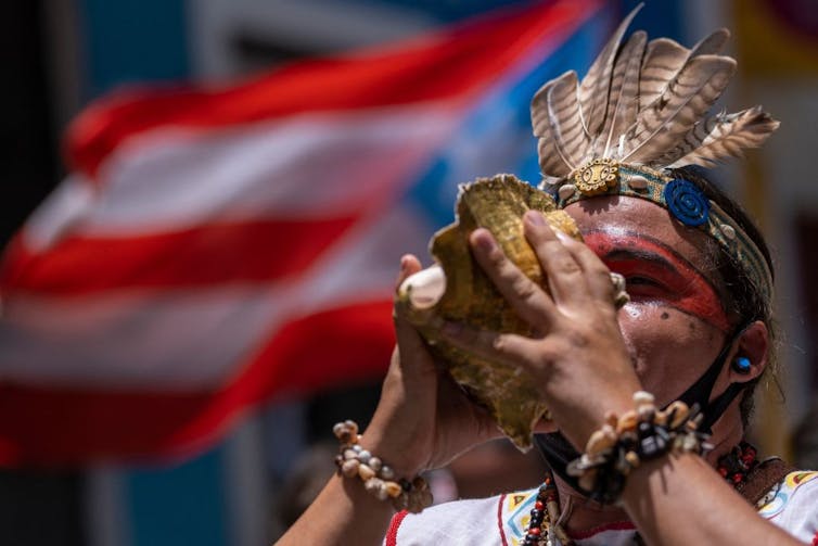 How both remix what it means to be Boricua A man in a feather headdress blows into a large shell, standing in front of a Puerto Rican flag.