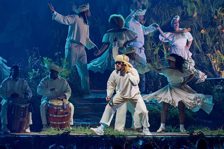 How both remix what it means to be Boricua A man in a white costume holds his hand up to his ear, looking off stage, as dancers in white outfits move behind him and beat drums.