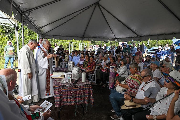 A few priests in white robes stand behind a table in an outdoor tent full of seated people.