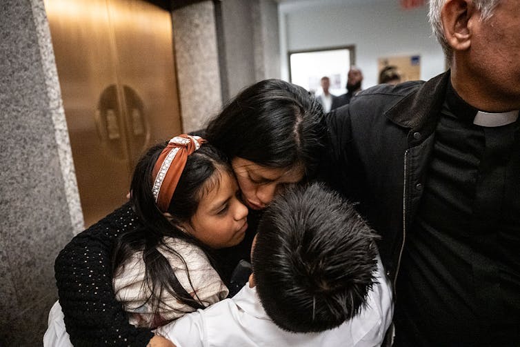 A man in a priest's collar, whose face is outside the frame, has his arm around an upset-looking woman and two children wearing white.