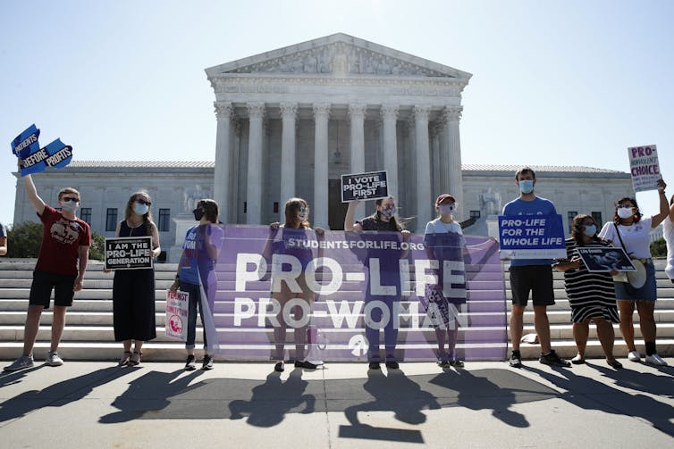 A line of protesters pose at the bottom of a long flight of steps, holding a sign that says 'Pro-life, pro-woman.'