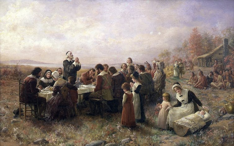 How the Plymouth Pilgrims took over Thanksgiving – and who history left behind A painting in muted colors of a small group of people in plain clothing seated around a table outside, with a log cabin in the background.